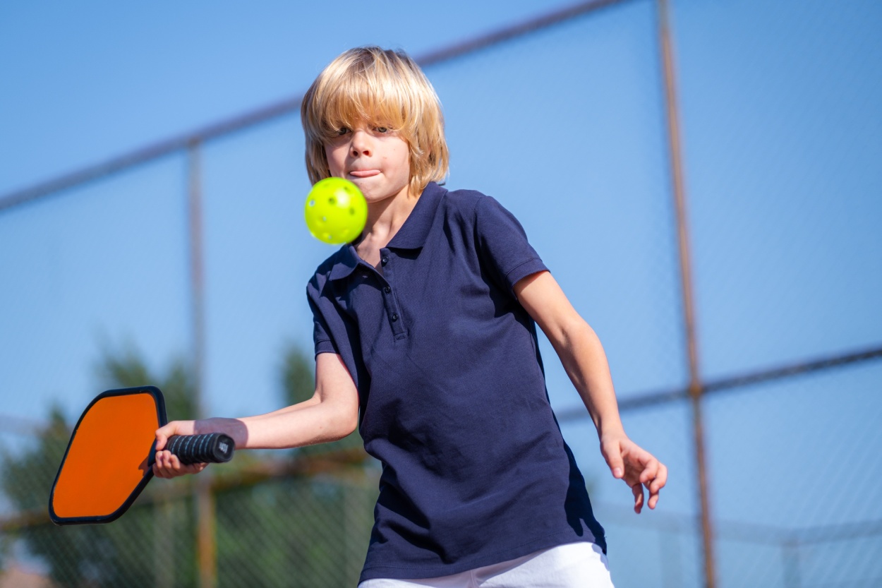 Happy blonde boy playing pickleball game, hitting pickleball yellow ball with paddle, outdoor sport leisure kids activity.